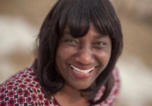 A smiling woman with long black hair, wearing a patterned red and white top, outdoors with a blurred background.