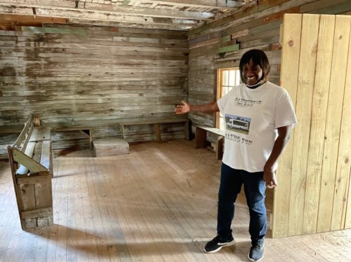 A man stands in a rustic, wooden room, gesturing towards the space with benches and a wooden door.
