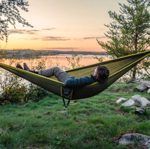 A person relaxes in a hammock by a serene lake at sunset, surrounded by trees and rocks.