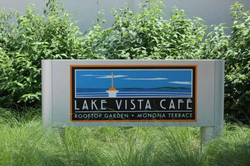 Sign for Lake Vista Café at Monona Terrace, featuring a colorful graphic of a lake and a tower. Surrounded by greenery.