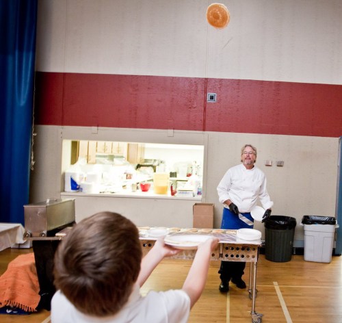 A chef tosses a pancake in the air while a child watches eagerly in a gymnasium setting.