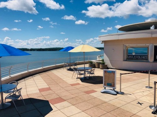 A sunny lakeside café patio with colorful umbrellas and tables overlooking the water and blue sky.