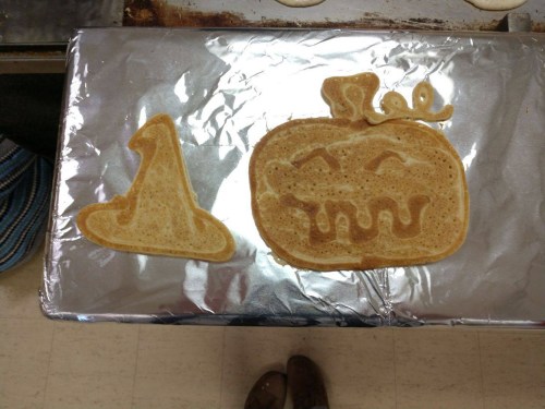 A silver tray with a witch hat and pumpkin-shaped pancakes, both decorated for Halloween.