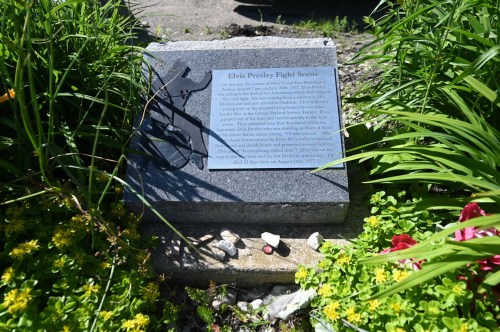 A stone plaque titled "Elvis Presley Flight Scene" surrounded by green plants and colorful flowers.