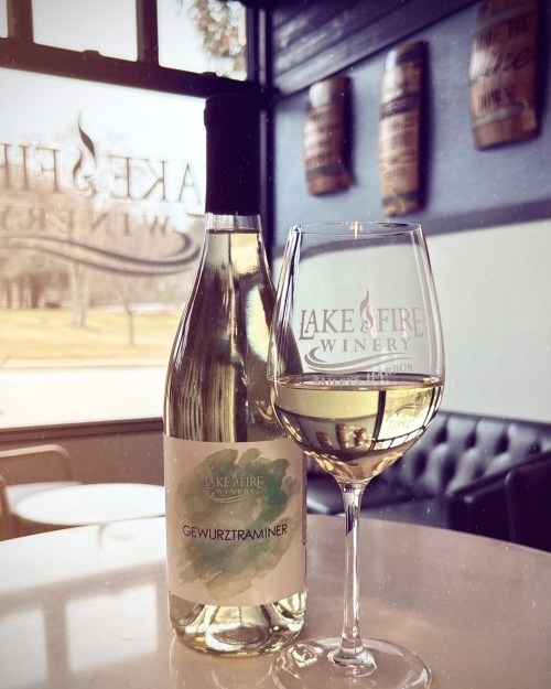 A bottle of Gewurztraminer wine next to a glass filled with the wine, set in a winery with wooden barrels in the background.