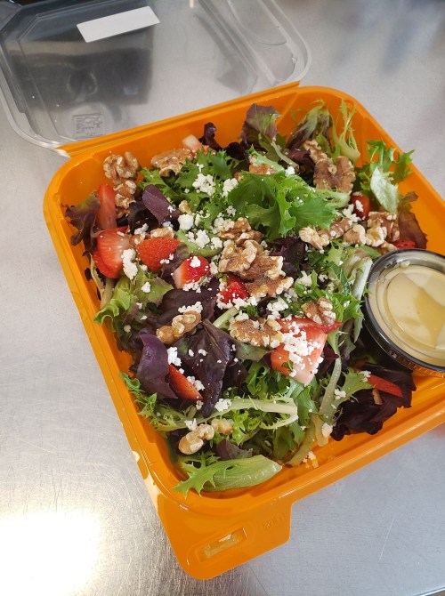 A colorful salad with mixed greens, strawberries, walnuts, feta cheese, and a small container of dressing in an orange box.