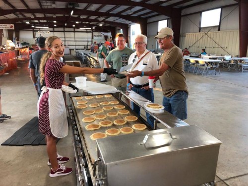 A young woman in an apron serves pancakes to three men at a large indoor event with tables and chairs in the background.