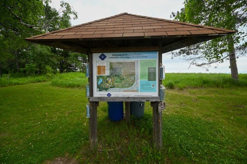 A wooden information kiosk with a map and welcome sign in a grassy area surrounded by trees.