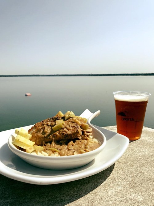 A bowl of savory dish with toppings and a cup of beer, set against a serene lake view under a clear sky.