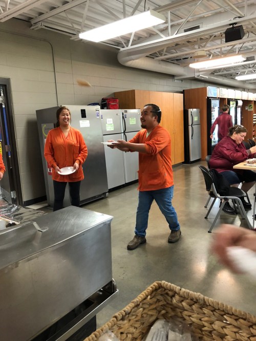 Two people in orange shirts laugh while one tosses a plate in a cafeteria setting, with others seated nearby.