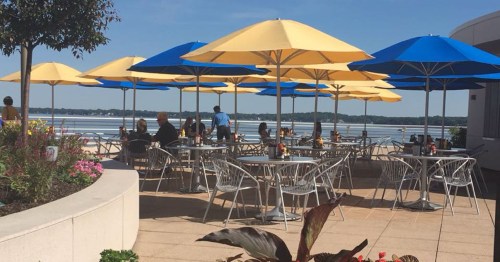 Outdoor dining area with yellow and blue umbrellas, tables, and a view of a lake on a sunny day.