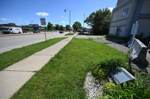 A sidewalk next to a road with a speed limit sign, grassy area, and a small flower bed under a clear blue sky.