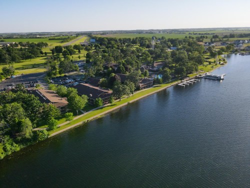 Aerial view of a lakeside area with green trees, a building, and a dock along the water's edge.