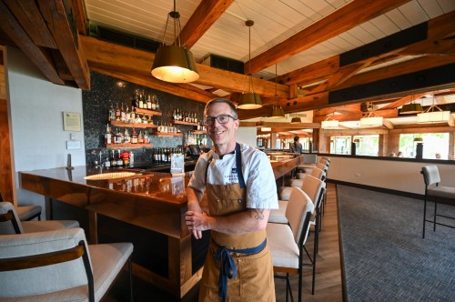A smiling chef stands at a stylish bar in a modern restaurant with wooden beams and a well-stocked shelf behind him.