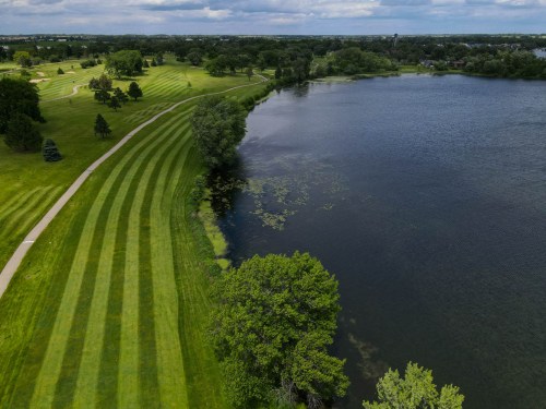 Aerial view of a lush green park with striped lawns beside a calm lake under a cloudy sky.