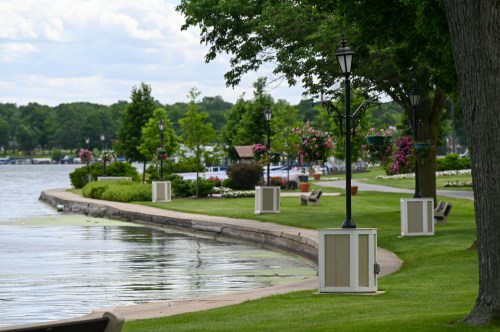 A scenic park along a lake, featuring green lawns, flower planters, benches, and decorative lamp posts.