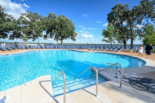 A clear blue pool surrounded by trees and lounge chairs, with a scenic view of the water and sky in the background.