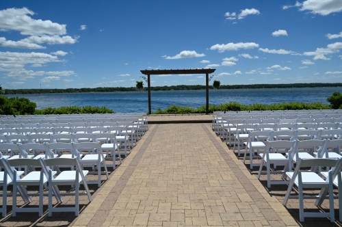 A scenic outdoor wedding setup with white chairs facing a wooden arch by a lake under a blue sky with clouds.