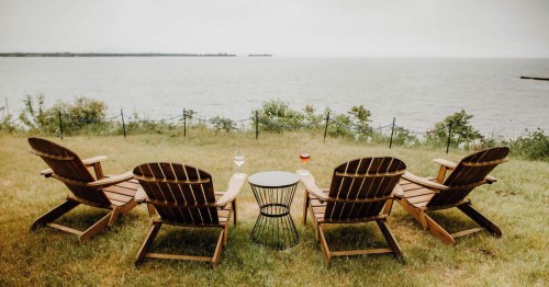 Four wooden lounge chairs facing a calm lake, with a small table between them, surrounded by green grass.