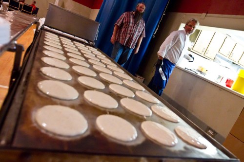 Two men stand near a large griddle filled with pancakes cooking in a kitchen setting.
