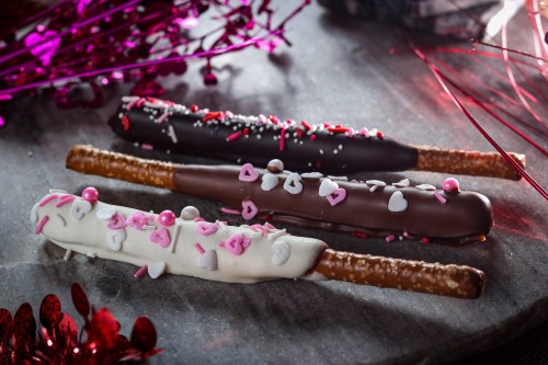 Three decorated chocolate-covered pretzel sticks with pink and white sprinkles on a marble surface, surrounded by festive decorations.