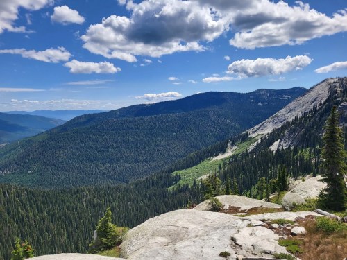 A panoramic view of lush green mountains under a blue sky with fluffy clouds, showcasing rocky terrain and dense forests.