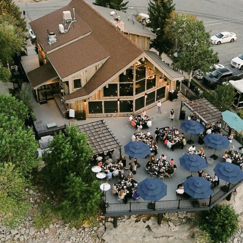 Aerial view of a rustic building with outdoor seating under umbrellas, surrounded by trees and parked cars.