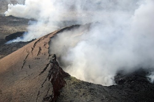 Aerial view of a volcanic crater with smoke rising from the edge and rocky terrain surrounding it.