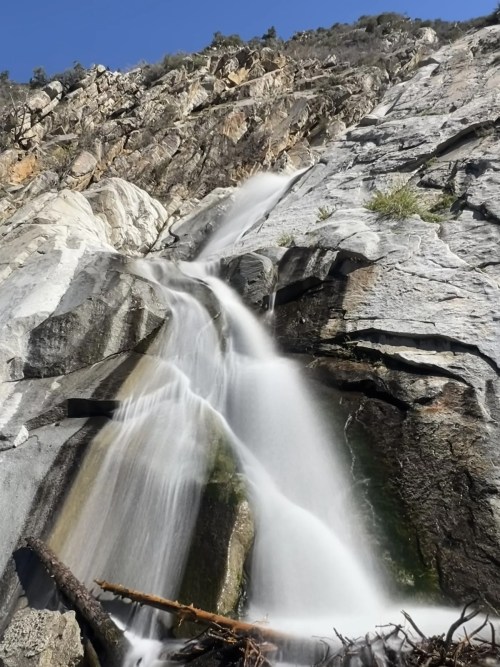 A cascading waterfall flows down a rocky cliff, surrounded by rugged terrain and clear blue skies.