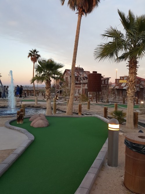 A mini-golf course with green turf, palm trees, and a water feature, set against a sunset sky.