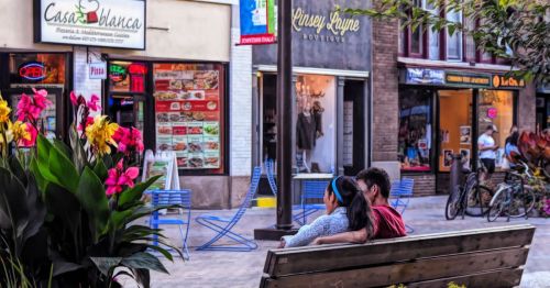 A couple sitting on a wooden bench in a vibrant outdoor shopping area in Ithaca, NY, featuring shops and a restaurant in the background.