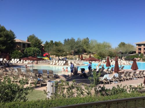 A sunny pool area with lounge chairs, umbrellas, and people enjoying the water, surrounded by greenery and buildings.