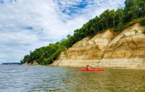 A person in a red kayak paddles along a calm river, with steep, tree-lined cliffs in the background under a cloudy sky.