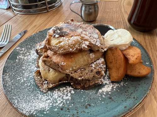 A plate of stacked pancakes dusted with powdered sugar, served with sliced apples and a dollop of cream.