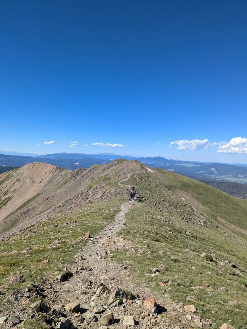 A winding trail leads over a mountain ridge under a clear blue sky, with hikers in the distance.