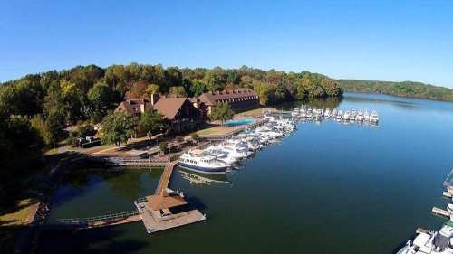 Aerial view of a marina with boats, surrounded by trees and a lodge by the water under a clear blue sky.