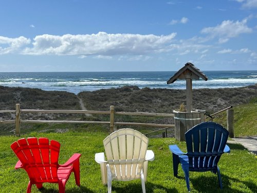 Three colorful adirondack chairs (red, white, blue) face a scenic ocean view with waves and a cloudy sky.