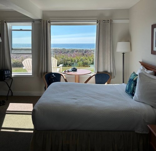 Cozy hotel room with a bed, chairs, and a scenic ocean view through large windows.