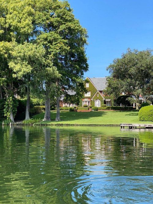 A serene lake view with lush trees and a large house in the background under a clear blue sky.