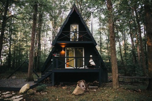 A black A-frame cabin nestled in a forest, surrounded by trees and featuring a porch and outdoor seating.