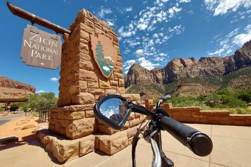 A bike handlebar in front of the Zion National Park entrance sign, with red rock formations and blue sky in the background.
