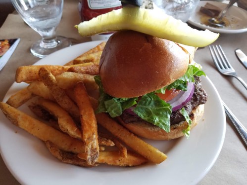 A juicy hamburger with lettuce, tomato, and onion, topped with a pickle, served with crispy fries on a white plate.
