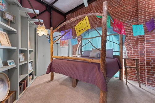 Cozy bedroom with a wooden bed, colorful papel picado decorations, and exposed brick walls. Shelves filled with framed art.