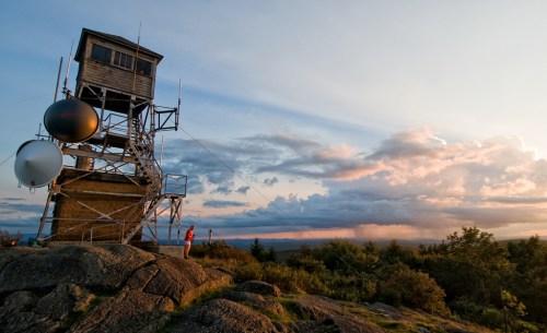 A person stands near a tall lookout tower on a rocky hill, with a colorful sunset and clouds in the background.
