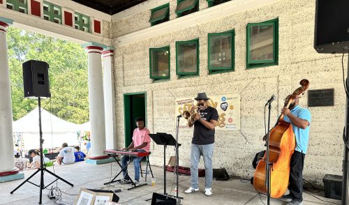 A live music performance featuring a keyboardist, a trumpeter, and a bassist on stage in a park setting.