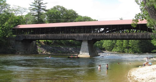 A covered wooden bridge spans a river, with people swimming and kayaking nearby, surrounded by lush greenery.