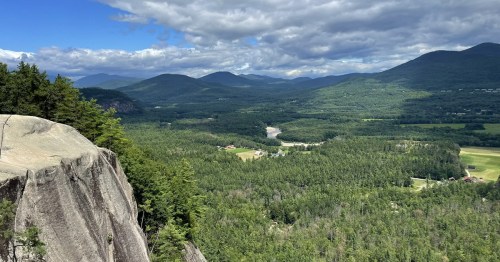 A panoramic view of lush green mountains and valleys under a partly cloudy sky, with a river winding through the landscape.