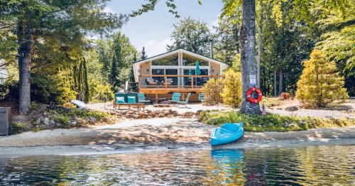 A serene lakeside cabin surrounded by trees, with a kayak on the water and outdoor seating on the shore.