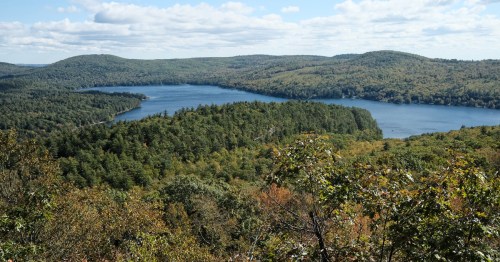 A scenic view of a lake surrounded by lush green forests and rolling hills under a partly cloudy sky.