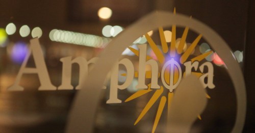 Close-up of a window with the word "Amphora" and a sunburst design, blurred city lights in the background.